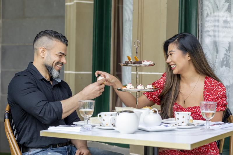a couple smiling at each other and enjoying high tea with Werribee Park Mansion behind them