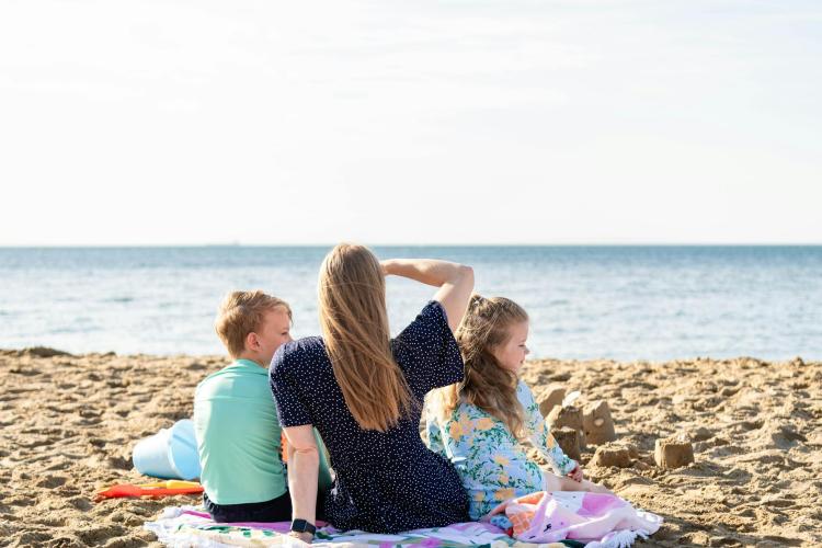 an adult and two children sitting on a towel on the sand looking out to the water