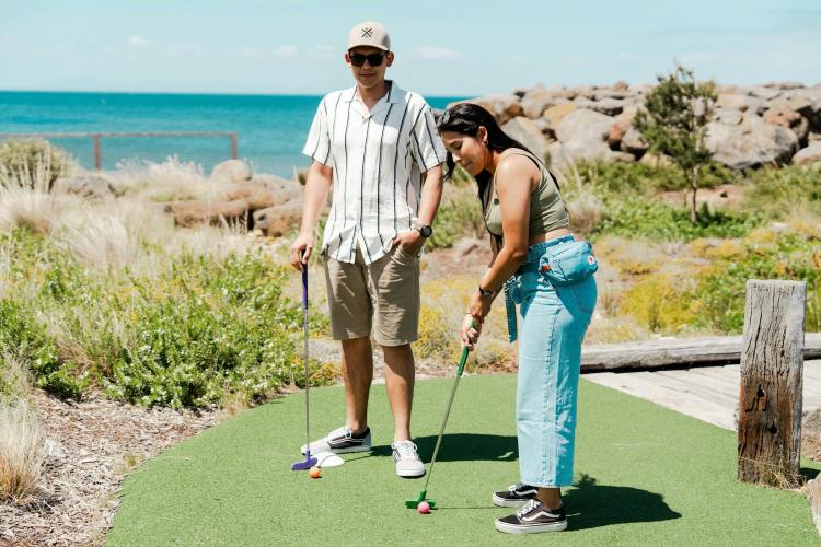 two adults playing mini golf in front of the ocean